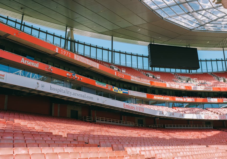 Inside the All England Club stadium in South Wimbledon, the image shows an empty football pitch with freshly mown grass arranged in a checkered pattern, surrounded by blue seating areas and tiered sta