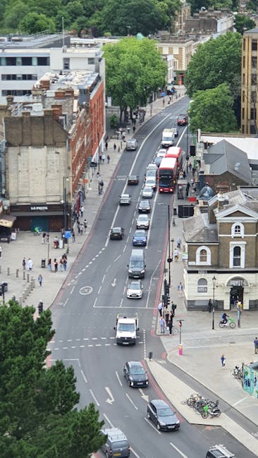 An aerial view of a residential street in South Wimbledon with a line of parked cars along the curb, including various small vehicles such as vans and sedans, while other cars are actively driving in both directions. The street is lined with trees and a mix of terraced brick houses and small commercial buildings, some with signage. Pedestrians are walking on the sidewalk, and a few bicycles are parked near the pavement. To the right, a small building with a pitched roof and a larger modern structure are visible. The scene depicts a typical urban street environment in South Wimbledon, appropriate for relocating or packing and moving activities, with overall lighting suggesting daytime. The environment provides context for home relocation services, highlighting the proximity of vehicles and pedestrians involved in logistics or furniture transport, as operated by Man with Van South Wimbledon.