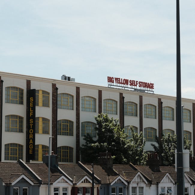 An exterior view of a multi-storey building with a sign reading 'BIG YELLOW SELF STORAGE' on the roof and a vertical sign on the side that says 'SELF STORAGE' in yellow letters. The building has cream walls accented with exposed brick columns and large arched windows with yellow frames. In front of the building, there are small residential houses with pitched roofs, chimneys, and front gardens. Green trees partially obscure the lower part of the building, and a lamppost is visible on the right side of the image. The scene is taken during daylight with a partly cloudy sky, reflecting typical urban surroundings associated with commercial and residential property, suitable for house removals and furniture transport services related to relocating or storage solutions, as provided by companies like Man with Van South Wimbledon.