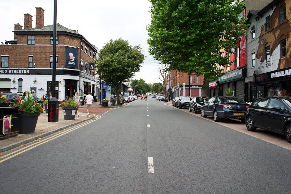 An aerial view of a residential street in South Wimbledon with a line of parked cars along the curb, including various small vehicles such as vans and sedans, while other cars are actively driving in both directions. The street is lined with trees and a mix of terraced brick houses and small commercial buildings, some with signage. Pedestrians are walking on the sidewalk, and a few bicycles are parked near the pavement. To the right, a small building with a pitched roof and a larger modern structure are visible. The scene depicts a typical urban street environment in South Wimbledon, appropriate for relocating or packing and moving activities, with overall lighting suggesting daytime. The environment provides context for home relocation services, highlighting the proximity of vehicles and pedestrians involved in logistics or furniture transport, as operated by Man with Van South Wimbledon.